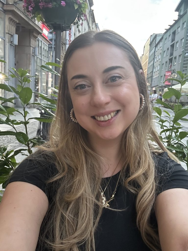 Photo of Beth smiling in a town centre with green foliage behind her. Beth has long blonde hair in a middle parting, hooped silver earrings and wearing a black top.